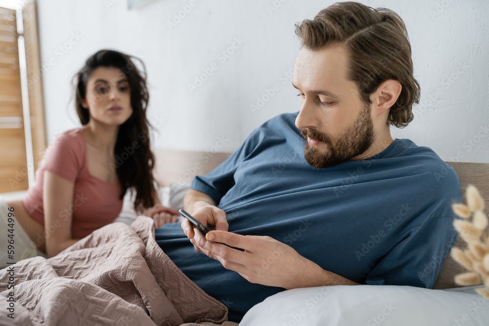 Obraz premium Bearded man using cellphone near blurred girlfriend on bed at home.