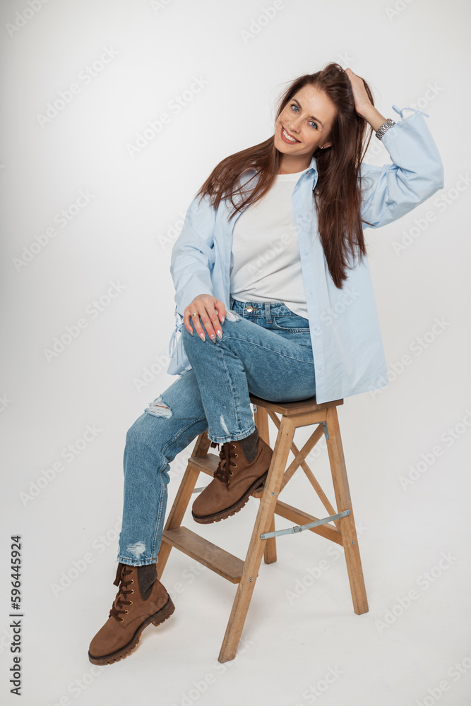 Cheerful beautiful young woman with a smile in fashion denim clothes with a stylish shirt, jeans and boots sits on a wooden chair on a white background Cheerful beautiful young woman with a smile in fashion denim clothes with a stylish shirt, jeans and boots sits on a wooden chair on a white background