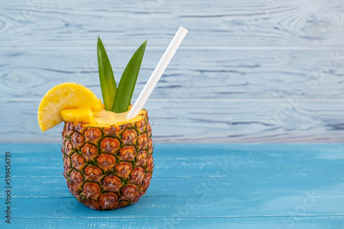 A horizontal photo featuring a pina colada cocktail served in a hollowed-out pineapple with pineapple wedges, a straw, and green leaves on a blue wooden background with copyspace on the right side of
