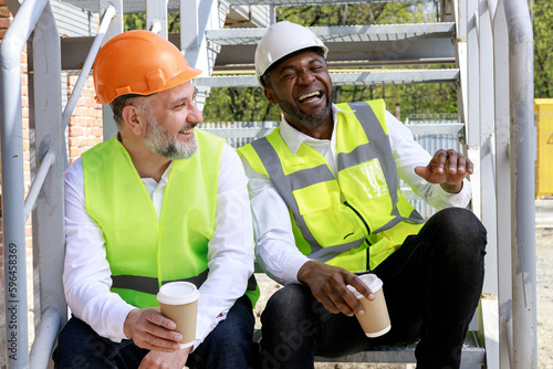 Smiling multicultural builders in hardhats, holding coffee sitting on stairs at construction site. Colleagues talking during enjoyable coffee break.