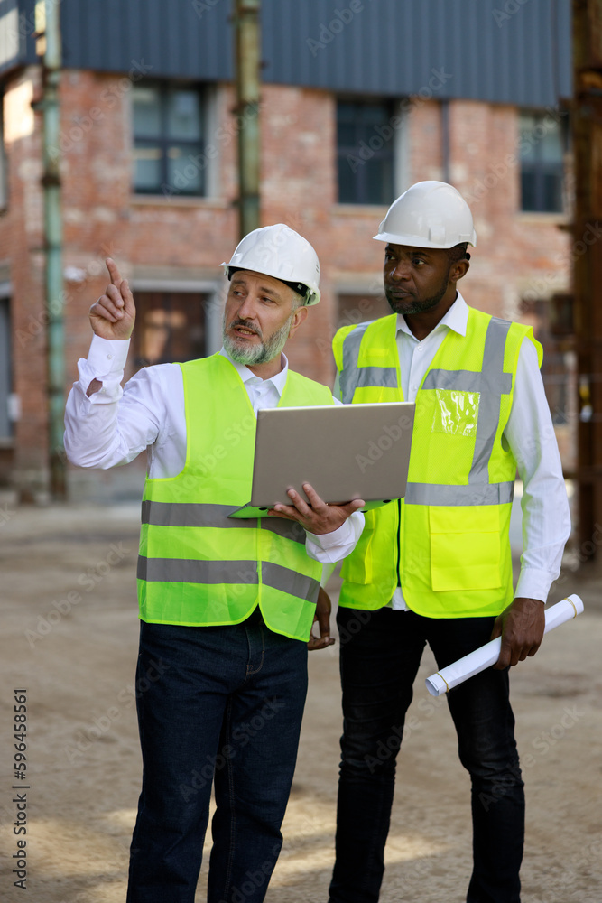 Builder with wireless laptop explaining all peculiarities of project to african american male architect at construction area. Concept of business meeting building process. Builder with wireless laptop explaining all peculiarities of project to african american male architect at construction area. Concept of business meeting building process.