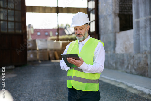 Architect in white hard hat and reflective vest standing, working on digital tablet at building site. High skilled mature builder using modern gadgets during construction work.