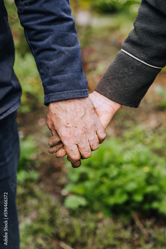 Elderly man and woman gardeners hold hands tightly while standing outdoors in a garden in nature. Close-up photography, eternal love concept, portrait of aging people.
