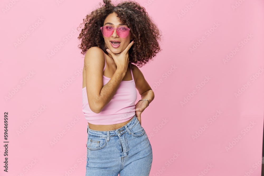 Happy woman with curly hair in a pink T-shirt and jeans on a pink background with sunglasses with a beautiful tan, DJ party, copy space
