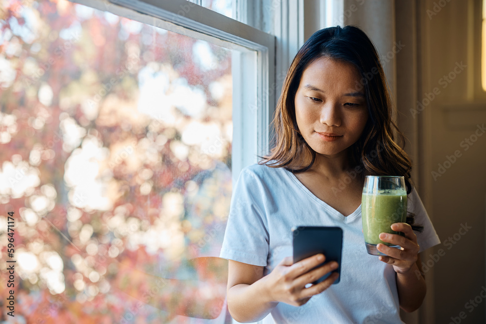 Young Asian woman drinks smoothie while using mobile phone by window.