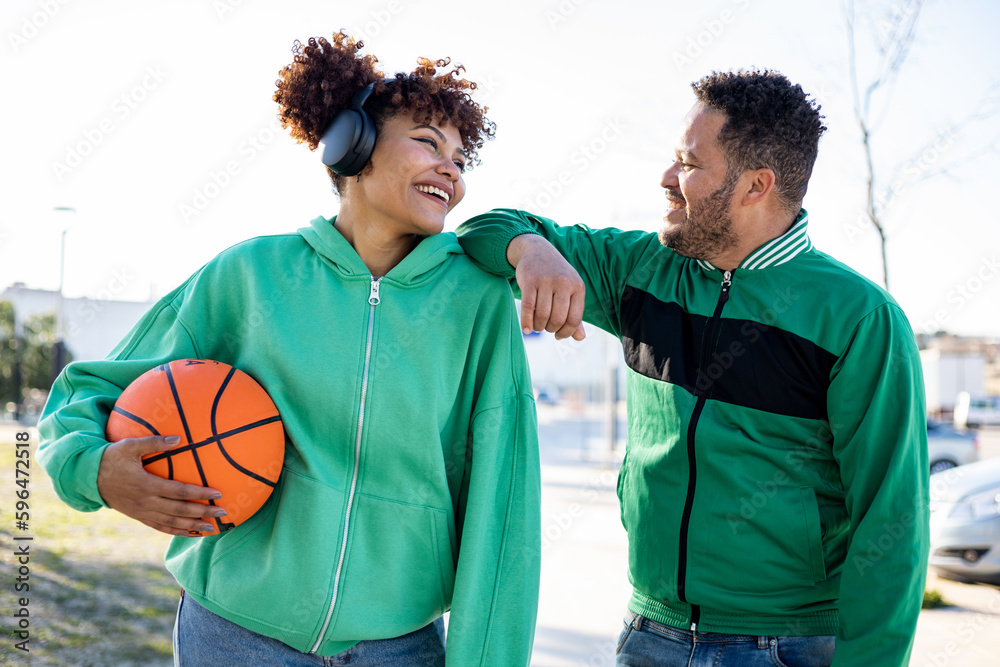 Two young men pose in the middle of the street in sportswear and a basketball. The girl with the afro hair looks happily at the chubby African boy who is leaning on her shoulder.