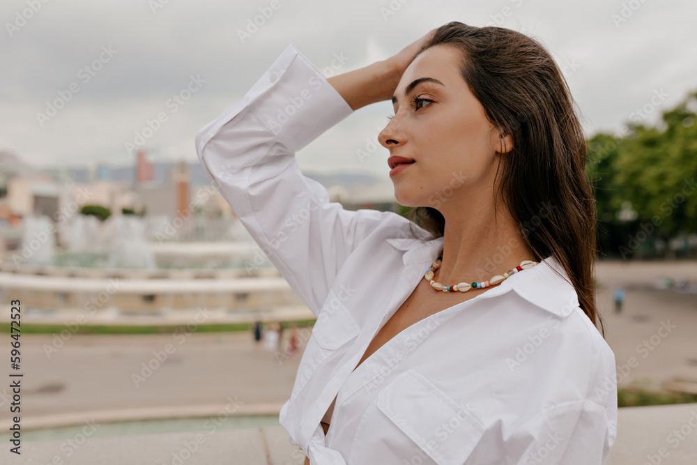 Close up profile photo of brunette woman with long dark hair wearing white shirt is touching her hair and looking staring with smile on background of city Close up profile photo of brunette woman with long dark hair wearing white shirt is touching her hair and looking staring with smile on background of city