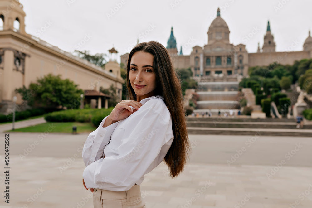 Spectacular young caucasian woman spending time outdoors and enjoying architecture of city. Brunette looks at camera, wears light shirt. Morning holiday concept