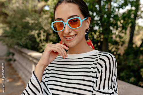 Cute young caucasian woman enjoying spending time outdoors in sunny weather. Brunette looks at camera, wears bright glasses and striped shirt. Relaxation concept