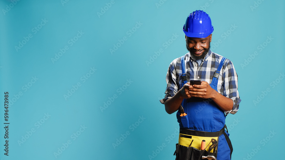 Construction worker texting messages on smartphone app, using social media browser in studio. Young craftsman browsing online website, wearing building overalls and safety helmet. Construction worker texting messages on smartphone app, using social media browser in studio. Young craftsman browsing online website, wearing building overalls and safety helmet.