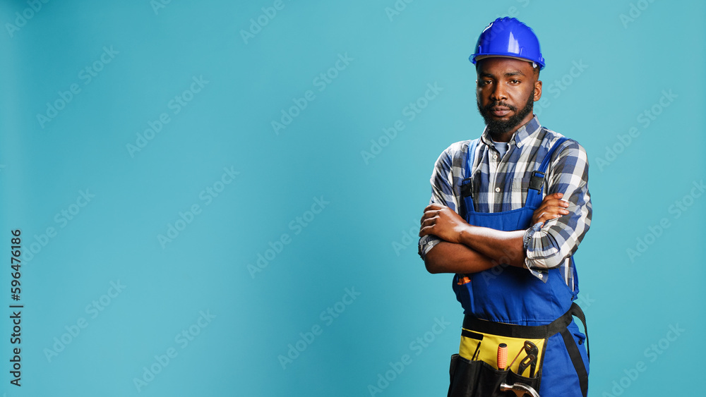 Cheerful smiling handyman standing over blue backdrop, wearing construction uniform and protective helmet. Professional carpenter repairman posing in studio, mechanical reconstruction.