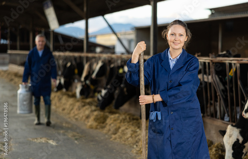 Portrait of positive woman farmer standing in cowshed, holding handle of working tool and smiling.