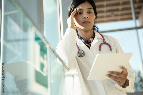 Serious Indian female doctor holding tablet, looking at camera. Sad intern in white coat