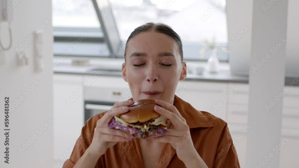 Junk food lover. Young happy woman biting big fresh tasty burger and licking her fingers, enjoying cheat meal at home