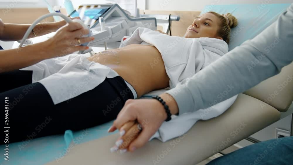 Beautiful pregnant lady undergoing ultrasound examination. Male hand takes his wife’s hand showing love and support. Beautiful pregnant lady undergoing ultrasound examination. Male hand takes his wife’s hand showing love and support.