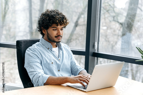 Smart successful confident indian or arabian man, company seo, IT specialist, HR manager, works on a laptop while sitting at a work desk in the office, looks at the laptop screen, smile