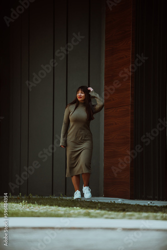 A confident and stylish plus-size woman walking by a modern building in an olive green dress, radiating beauty, empowerment, and self-love.