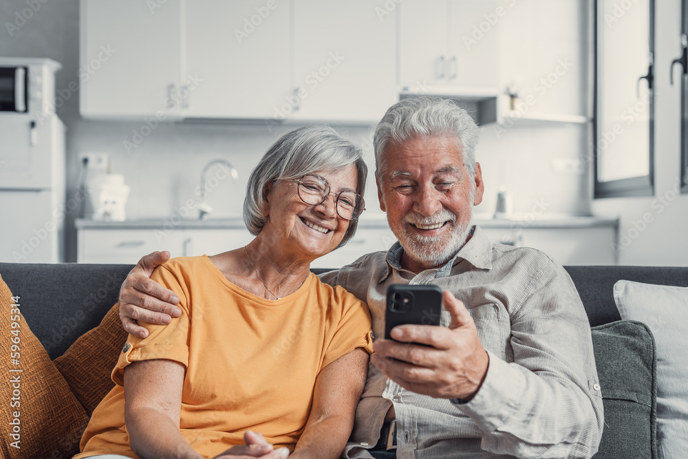 Happy mature family, wife and husband using phone together at home, smiling middle aged man and woman sitting on couch, using mobile device apps, watching video in social network, surfing internet.