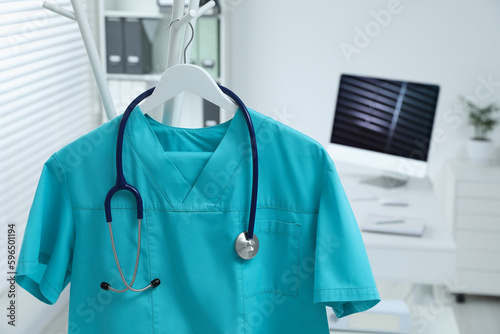 Turquoise medical uniform and stethoscope hanging on rack in clinic, closeup. Space for text