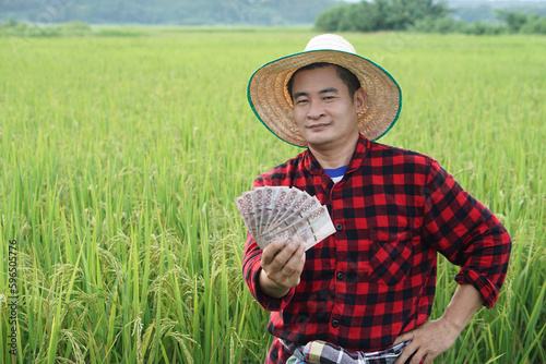 Asian farmer man is at paddy field, wears hat and red plaid shirt, hold Thai banknote money. Concept : Farmer happy to get profit, income, agriculture supporting money.      