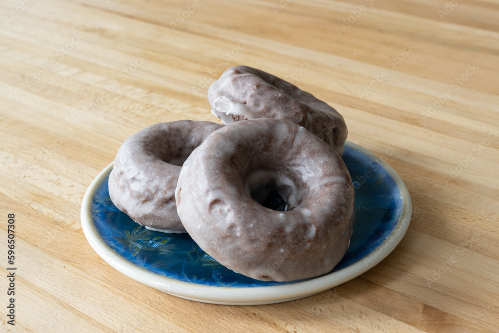 Multiple homemade dark chocolate doughnuts covered in a sugar vanilla glaze. The sweet baked pastries are on a small blue plate and oak wood table. Multiple homemade dark chocolate doughnuts covered in a sugar vanilla glaze. The sweet baked pastries are on a small blue plate and oak wood table.