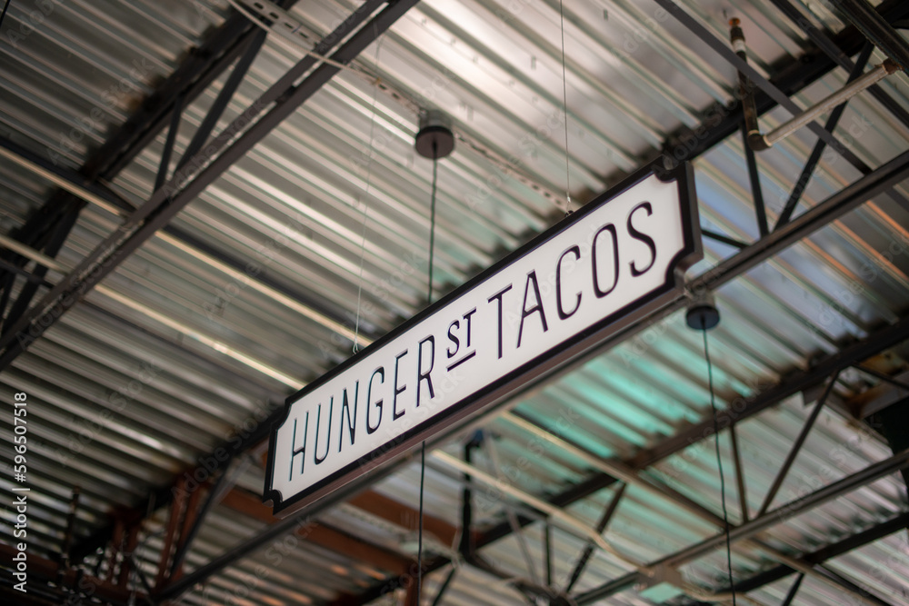 A white with black lettering sign hanging from the ceiling of a market. The sign spells hunger street tacos. The interior of the industrial warehouse ceiling is silver metal with a black grid system.