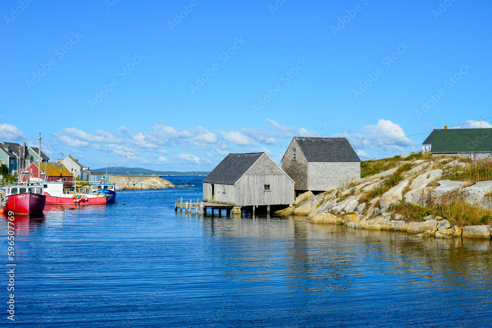 Scenic Peggy's Cove, Nova Scotia, is a quaint fishing village on the edge of a rocky coastline. The storage buildings are grey and weathered.  The summer sky and water are blue with white clouds. 
