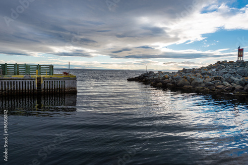 A wooden wharf with yellow and green boards. The dock is empty. The cove has a rocky breakwater with a navigational sign on top of the point. The ocean is blue and calm with a ripple from the wind. 