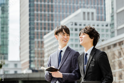 オフィス街で会話しながら歩く20代と30代のスーツ姿のビジネスマンの男性2人のチーム