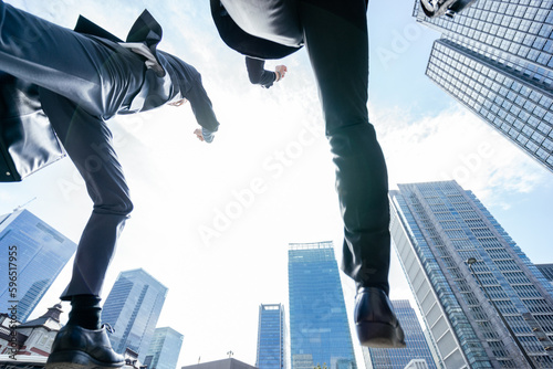 ビル群に囲まれたビジネス街でローアングルから撮影したジャンプとダッシュするスーツの2人の男性 photo