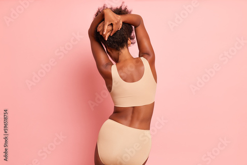 Shot of dark-skinned girl from back in underwear on a pink background, wears body-colour panties and a top, stands with her hands up to her hair, graceful pose and beautiful body concept, copy space