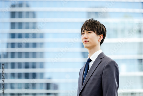 オフィスビルのガラスの建物の前で真面目な表情で遠くを見つめる20代のスーツを着た新入社員の男性