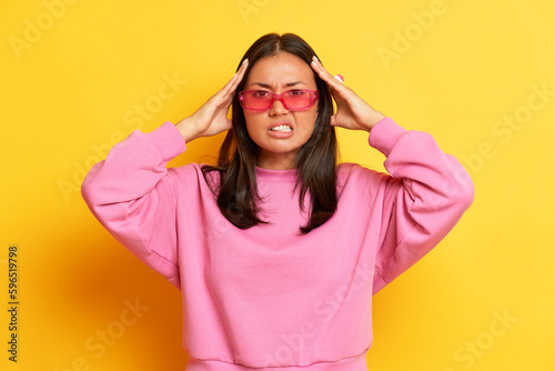 Serious brunette woman in pink sweatshirt dissatisfied with something, holds her hands on her head, stands on yellow studio backdrop, copy space, high quality photo © South House Studio