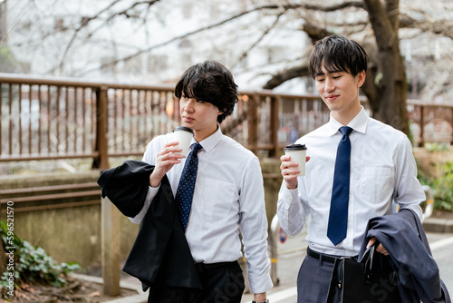 東京の中目黒の川沿いの緑道をテイクアウトドリンクを持って並んであるくスーツを脱いでシャツ姿の男性2人