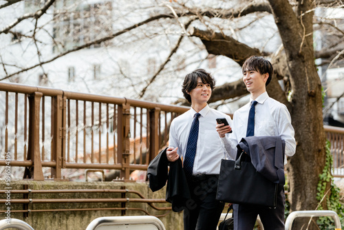 桜の緑道のある街並みをスマートフォンを持って会話しながら笑顔で並んで歩く2人の若いビジネスマンの男性