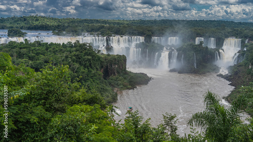 An impressive cascade of Iguazu Waterfalls falls down. A tourist boat is floating on the river. Lush tropical vegetation all around.Clouds in the sky. Brazil