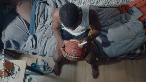 Young black basketball player sitting on couch at home, writing autograph on ball with marker pen. Top down shot