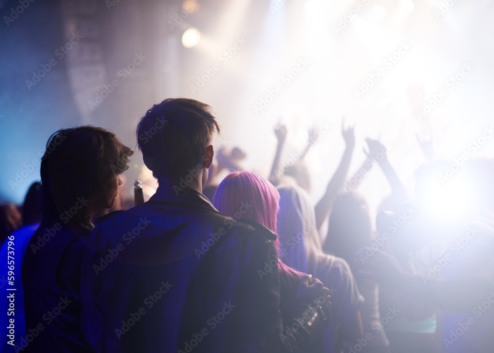 Fototapeta premium Dance, rock and friends in crowd at music festival from back, watching live band performance on stage. Audience in arena, lights and group of people at party, fans of musician at concert together.