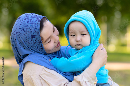 Cute young islamic kazakh woman hugging her little crying son, newborn boy. Yappy asian mother in hijab soothes, holding baby in hands outdoors. Family concept photo