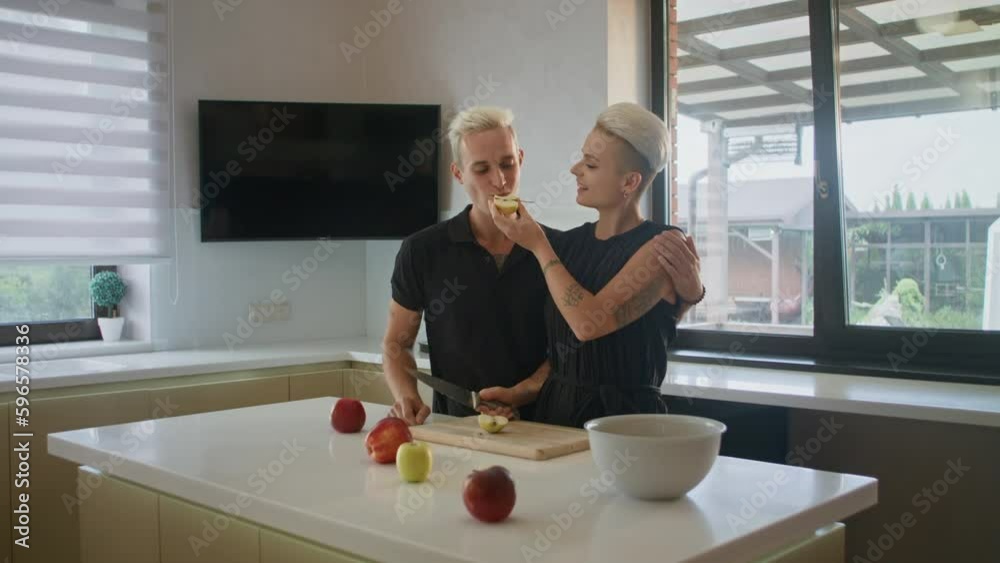A loving couple stands at the table. They both have similar dyed short blond hair. The girl cuts a green apple in half and gives her boyfriend a bite. She takes a bite after. They seem satisfied. A loving couple stands at the table. They both have similar dyed short blond hair. The girl cuts a green apple in half and gives her boyfriend a bite. She takes a bite after. They seem satisfied.