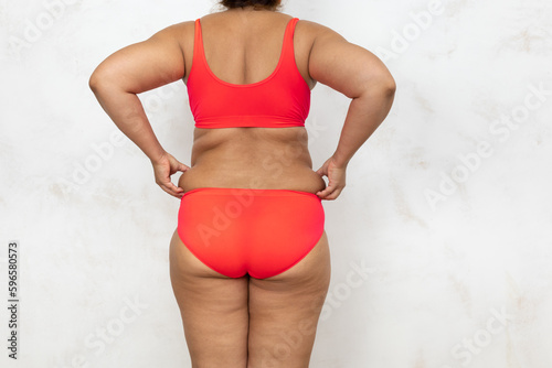 Photo of half of female cellulite body with back, free copy space, white background. Bare woman in red underwear pinch fat folds by hands. Overweight people, weight loss, self acceptance.