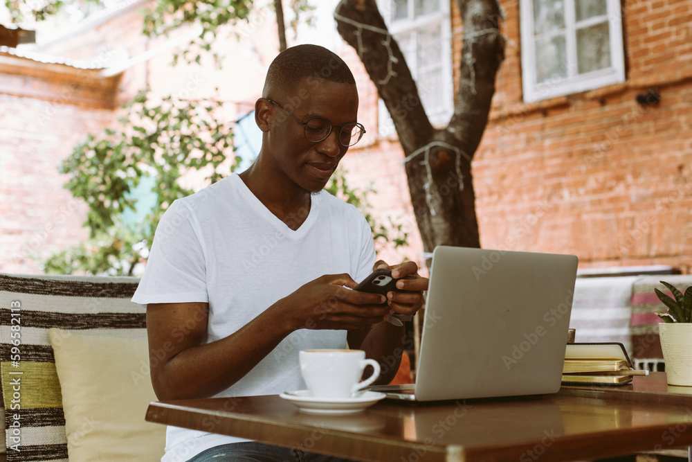 Handsome afro american man with glasses sitting at table in cafe with cup of coffee on summer veranda, holding and looking at smartphone. Working online on laptop, business man, freelance. Handsome afro american man with glasses sitting at table in cafe with cup of coffee on summer veranda, holding and looking at smartphone. Working online on laptop, business man, freelance.
