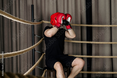 Big guy sits at the corner of a boxing ring, wearing red head guard helmet and gloves and drinking from a sport bottle