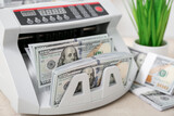 Modern cash counting machine with dollar banknotes and grass on table in room, closeup