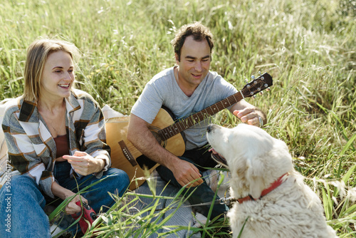 Happy caucasian couple with their labrador dog sitting on the lawn in summer nature with guitar.