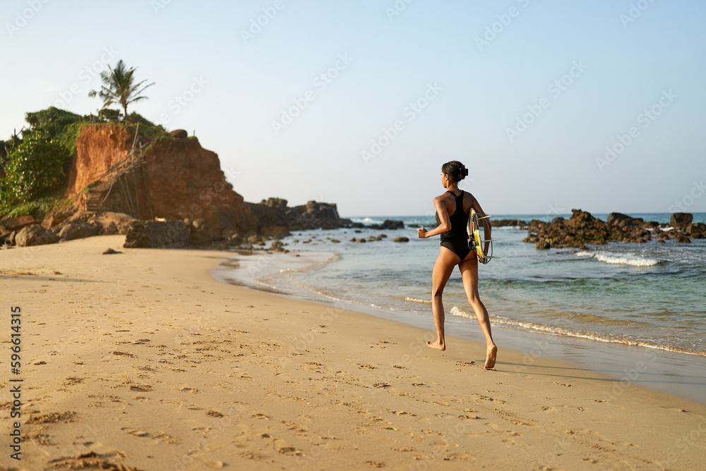 African american woman running with surfboard on ocean beach. Black female surfer with surf board. Pretty multiethnic girl goes on surfing session on tropical location at sunny sunrise.