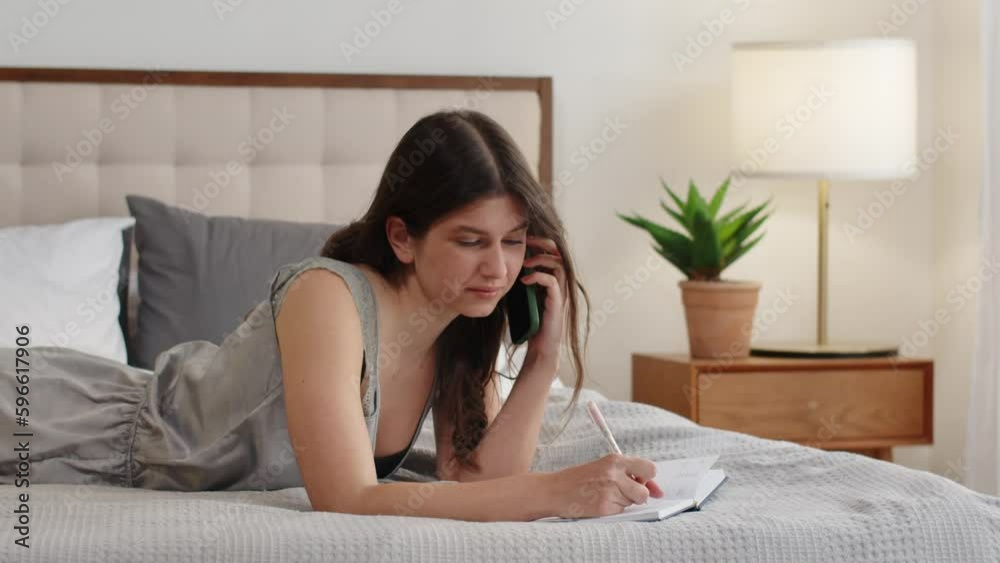A beautiful brunette is talking on the phone, lying on the bed and writing something in a notebook. A young woman discusses weekend plans with her friend on the phone. A beautiful brunette is talking on the phone, lying on the bed and writing something in a notebook. A young woman discusses weekend plans with her friend on the phone.