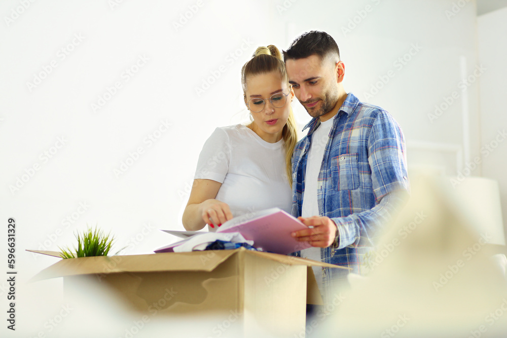A happy middle-aged Caucasian husband and wife standing in their new home and taking out framed photo album out of cardboard box, chatting animatedly and laughing A happy middle-aged Caucasian husband and wife standing in their new home and taking out framed photo album out of cardboard box, chatting animatedly and laughing