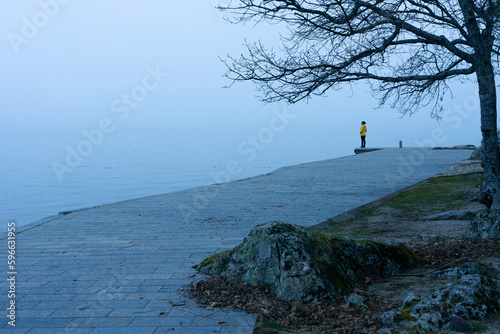 Woman with a yellow jacket looking at the horizon on a pier on a very foggy day at Lake Sanabria. Zamora, Spain.