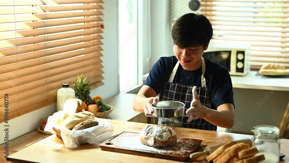 Happy millennial man wearing apron sifting flour from sieve on dough, making bread in kitchen Happy millennial man wearing apron sifting flour from sieve on dough, making bread in kitchen